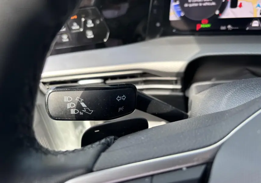 Détail du commodo gauche derrière le volant d’une Volkswagen Golf blanche 2024, avec tableau de bord numérique en arrière-plan.