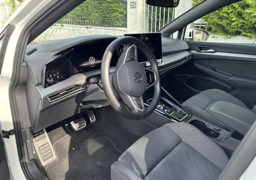 Vue intérieure côté conducteur de la Volkswagen Golf blanche 2024, mettant en valeur le tableau de bord digital et le volant multifonction.
