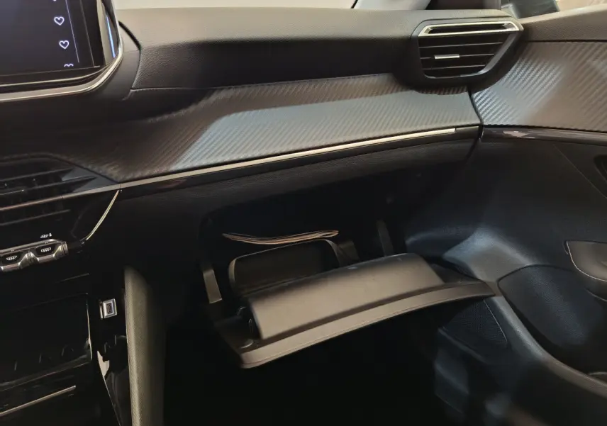 Intérieur de la Peugeot 208 PureTech 100 S&S Allure 2025, vue du coffre à gants ouvert côté passager, tableau de bord gris clair.