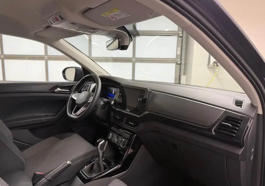 Vue intérieure côté conducteur du Volkswagen T-Cross 2025, avec tableau de bord noir et sièges gris foncé.