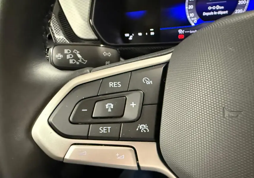 Gros plan sur les commandes au volant du Volkswagen T-Cross 2025, avec boutons de régulateur et affichage numérique bleu.