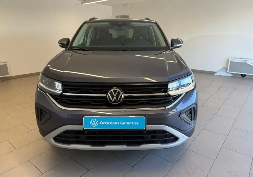 Vue avant d'un Volkswagen T-Cross 2025 gris cendre, avec calandre noire et logo VW centré, en intérieur showroom.