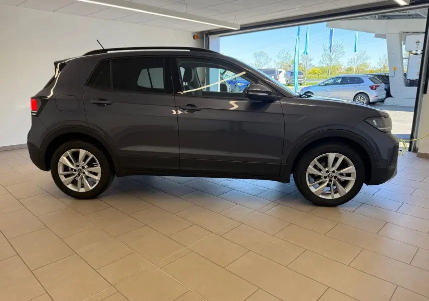 Profil côté gauche du Volkswagen T-Cross gris cendre 2025, version 1.0 TSI VW Edition, en intérieur lumineux.