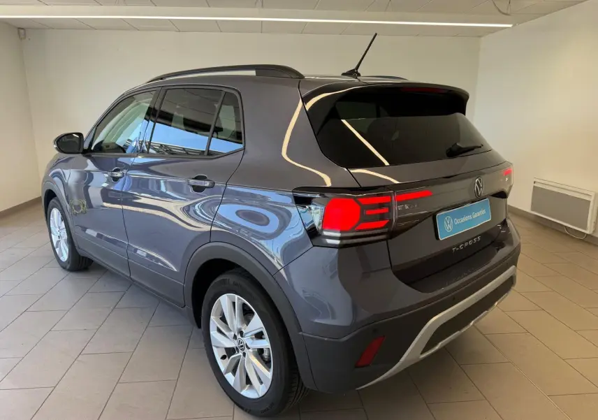 Volkswagen T-Cross gris cendre vu en 3/4 arrière droit, avec feux arrière allumés et jantes argentées.