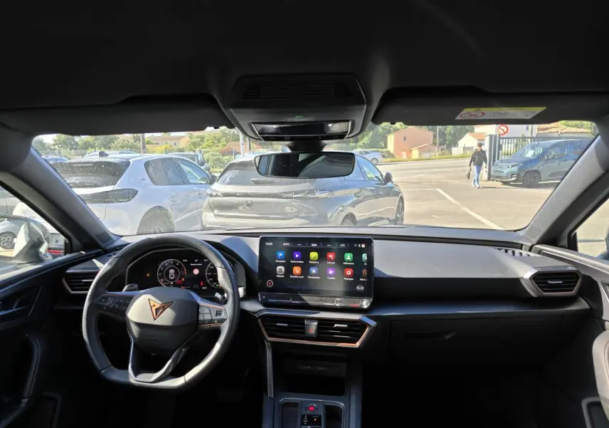 Vue intérieure du tableau de bord et volant Cupra d'une Leon Sportstourer 2024, avec écran tactile central allumé.