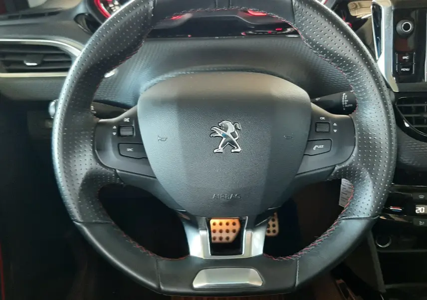 Volant en cuir perforé avec surpiqûres rouges et logo Peugeot, tableau de bord et pédales sportives visibles en arrière-plan.