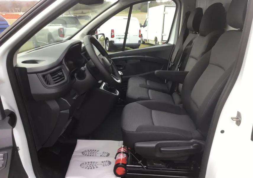 Vue intérieure côté conducteur du Renault Trafic blanc, montrant les sièges noirs et le tableau de bord avec volant et levier de vitesse.