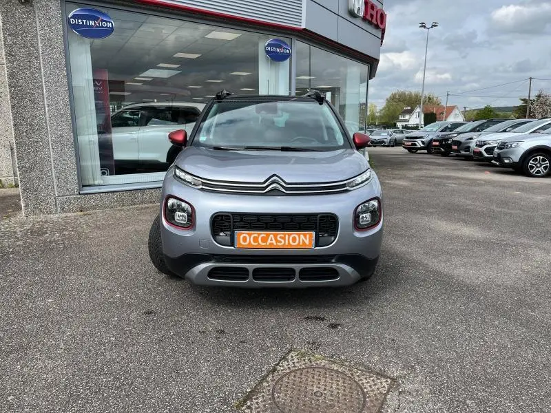 Citroën C3 Aircross gris acier vue de face avec barres de toit noires et rétroviseurs rouges, devant un concessionnaire.