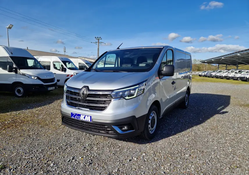 Renault Trafic Fourgon gris Highland vu en 3/4 avant droit sur parking avec plusieurs autres utilitaires blancs.