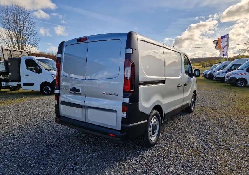 Vue 3/4 arrière droite d’un Renault Trafic Fourgon gris Highland garé sur un sol gravillonné avec plusieurs autres utilitaires en arrière-plan.