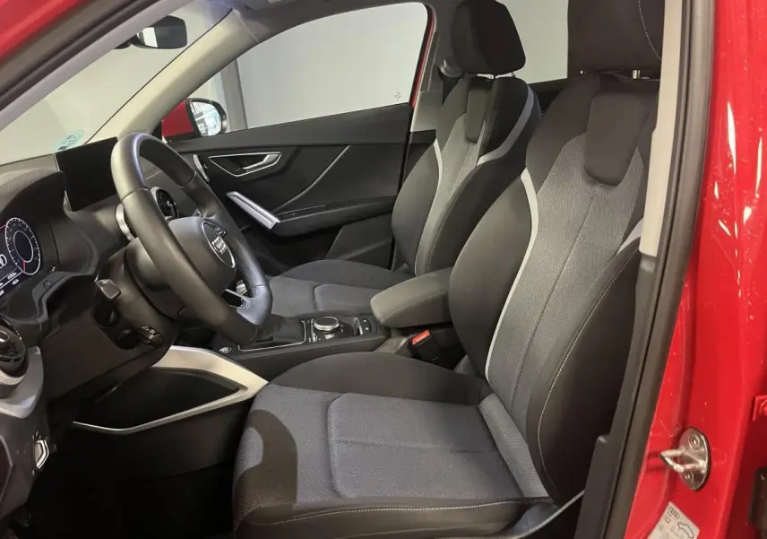 Vue intérieure côté conducteur de l'Audi Q2 rouge 2024, sièges sport noirs avec surpiqûres grises et volant cuir multifonction.