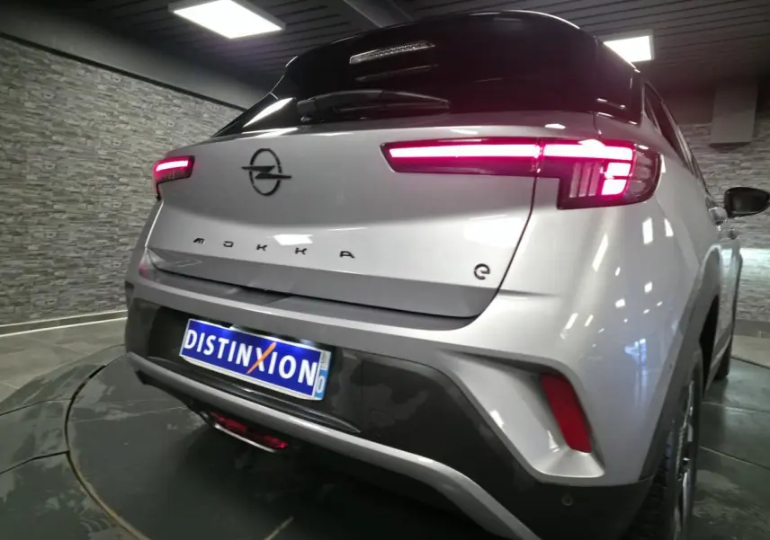 Vue 3/4 arrière droite d'un Opel Mokka-e gris avec feux arrière LED allumés et logo noir sur coffre.