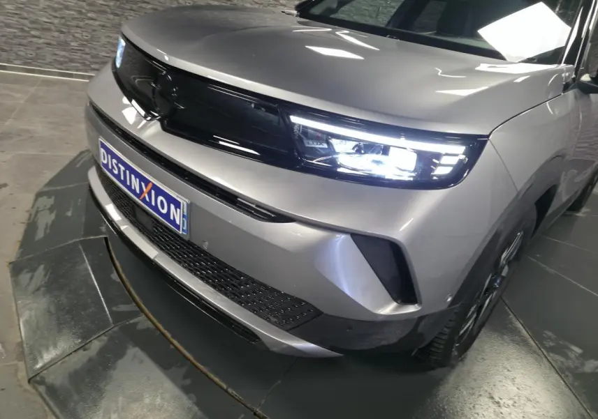 Vue 3/4 avant droit de l'Opel Mokka-e gris métallisé avec calandre noire et phares Matrix LED allumés.