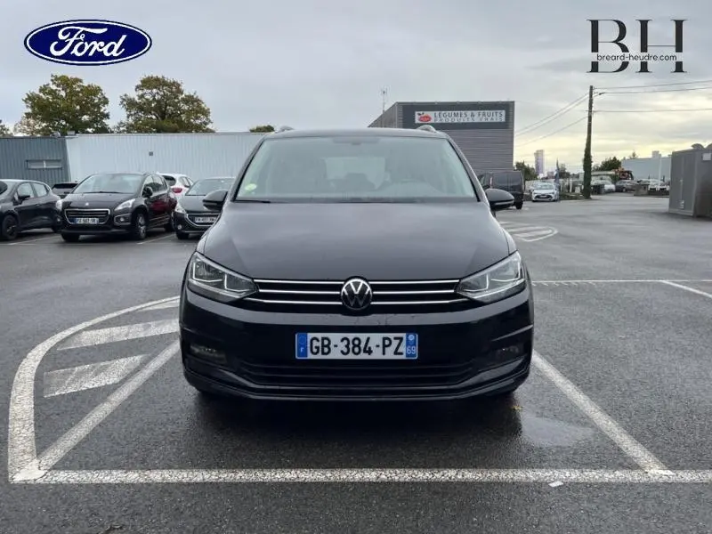 Vue de face d'un Volkswagen Touran noir intense nacré garé sur un parking sous un ciel nuageux.