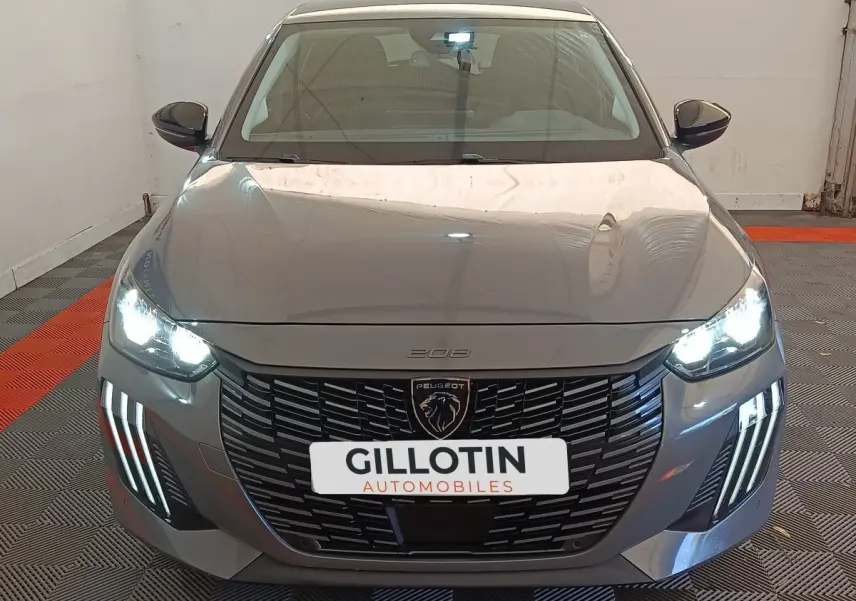 Vue avant d'une Peugeot 208 gris foncé avec phares allumés et calandre noire distinctive en intérieur showroom.