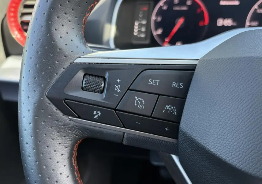 Gros plan sur les commandes à gauche du volant en cuir perforé noir avec surpiqûres rouges du SEAT Ibiza blanc Candy 2025.