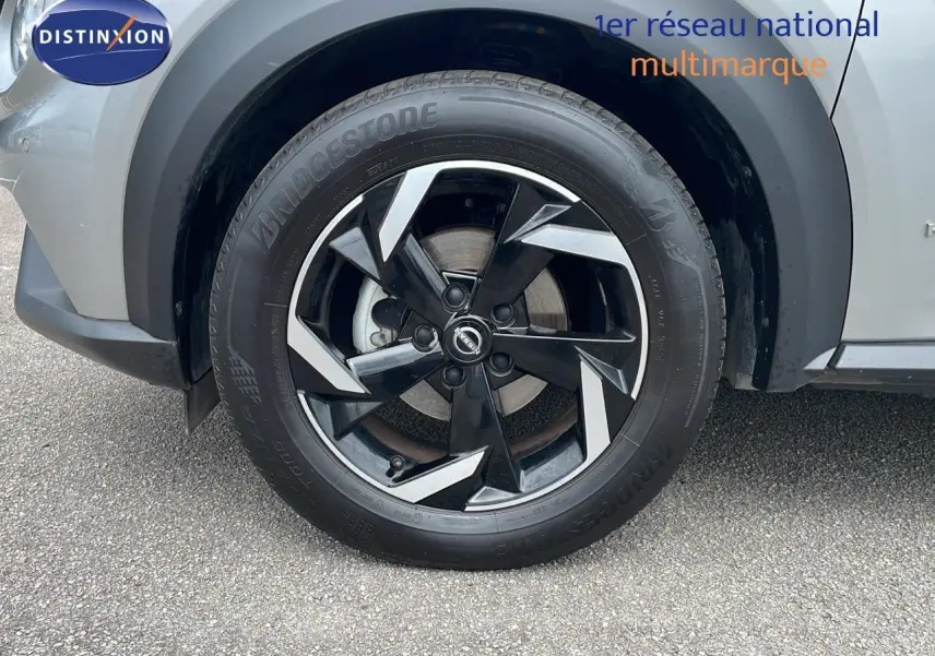 Gros plan sur la roue avant gauche du Nissan Juke Hybrid 143ch Business+ gris perle métal avec jante noire et argent.