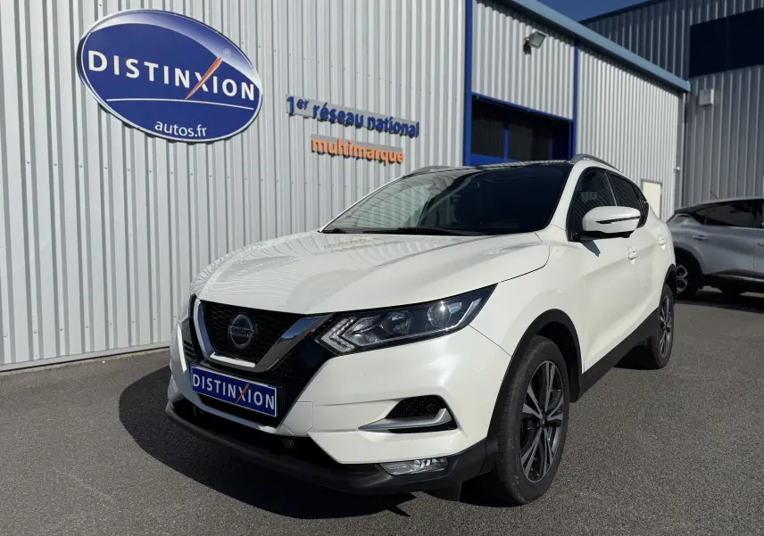 Nissan Qashqai blanc en 3/4 avant droit, avec jantes noires et calandre chromée, stationné devant un bâtiment industriel.