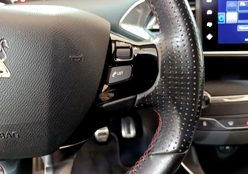 Gros plan sur le volant cuir noir perforé avec surpiqûres rouges et partie droite du tableau de bord tactile d’une Peugeot 308 gris foncé.