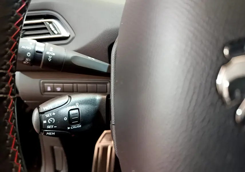Gros plan sur la commande de régulateur de vitesse à gauche du volant cuir noir avec surpiqûres rouges dans un Peugeot 308 gris foncé.