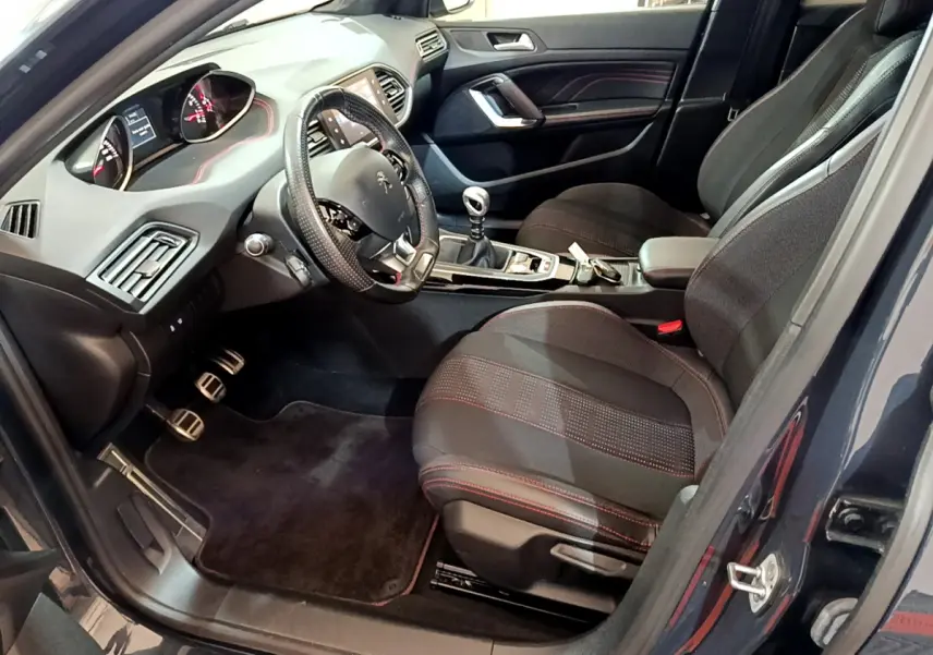 Intérieur avant droit de Peugeot 308 gris foncé, volant cuir, sièges tissu noirs avec surpiqûres rouges, boîte manuelle visible.