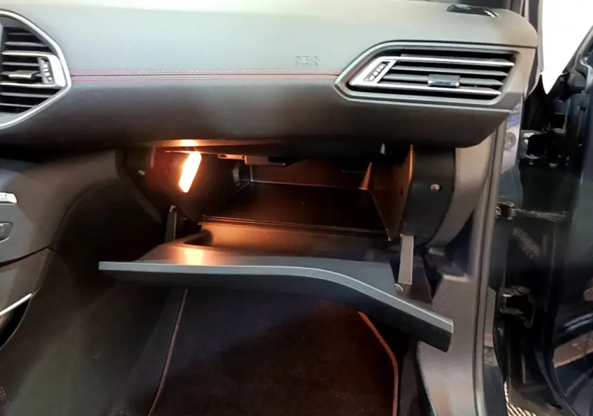 Gros plan sur la boîte à gants ouverte côté passager d’une Peugeot 308 gris foncé avec éclairage intérieur.