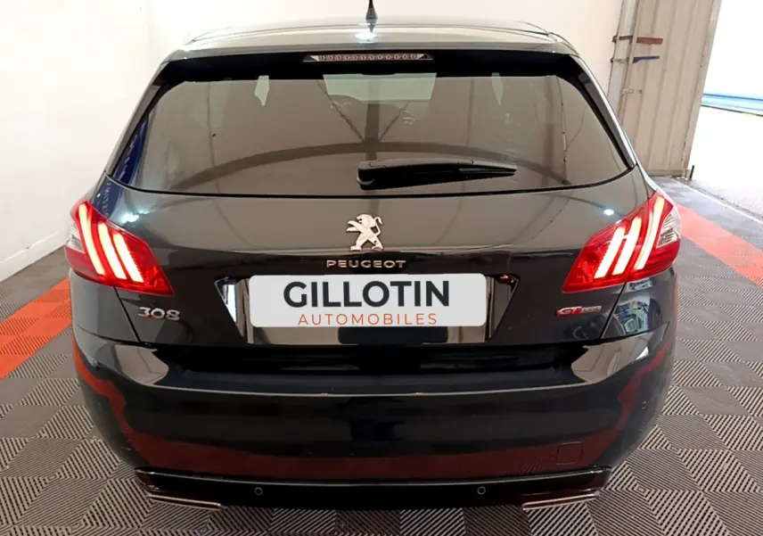Vue arrière d'une Peugeot 308 gris foncé avec feux allumés et logo GT Line visible sur un sol à damier.