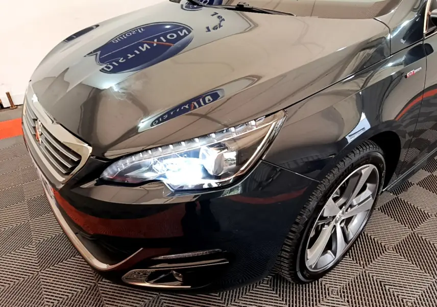 Vue 3/4 avant droit d'une Peugeot 308 gris foncé avec phares allumés et jantes alliage visibles.