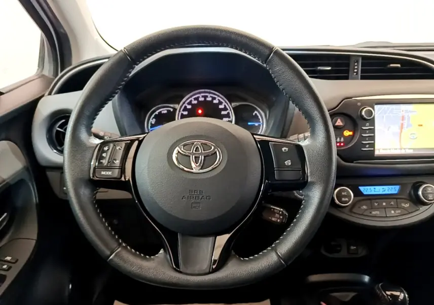 Vue intérieure centrée sur le volant cuir et alu de la Toyota Yaris blanche, avec tableau de bord et écran tactile visible.