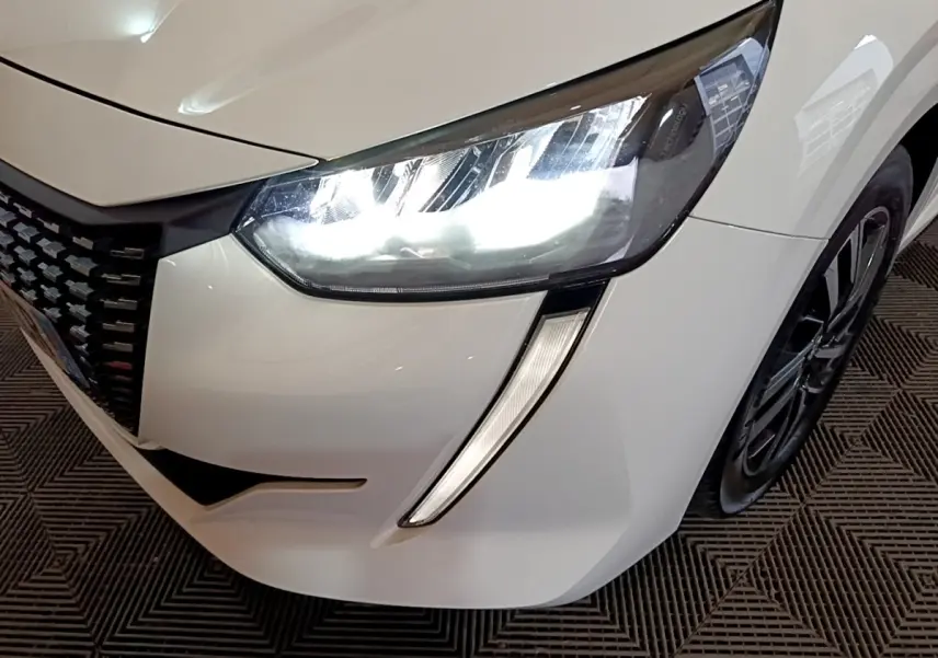 Gros plan sur l'avant droit blanc d'une Peugeot 208 2021, mettant en valeur le phare LED allumé et la calandre noire.