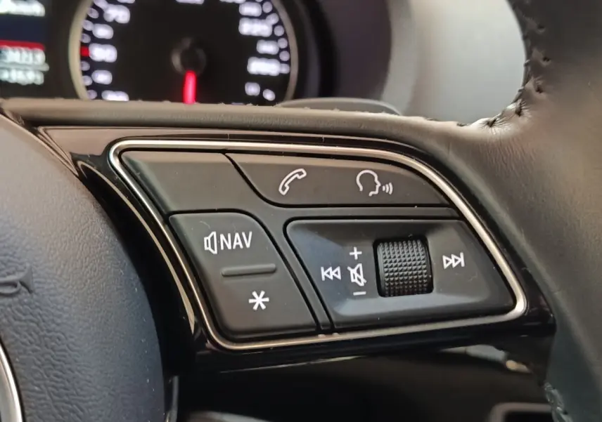 Gros plan sur les commandes multifonctions du volant cuir de l'Audi A3 blanc, avec tableau de bord flou en arrière-plan.