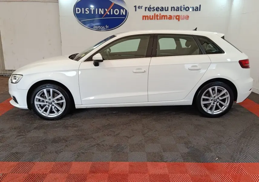 Profil latéral droit d'une Audi A3 blanche 35 TDI 150 S tronic Business Line sur sol noir et rouge en intérieur showroom.