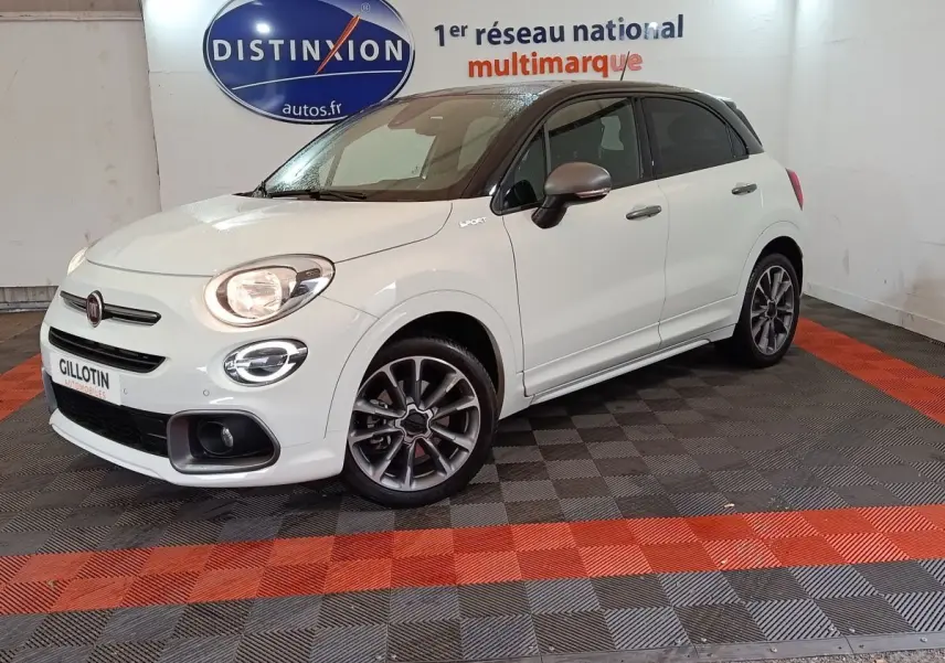 Fiat 500X blanc à toit noir en 3/4 avant droit, avec jantes sport et feux allumés dans un showroom.