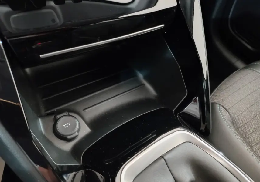 Gros plan sur la console centrale noire avec prise 12V et partie du siège tissu gris de la Peugeot 208 PureTech 100.