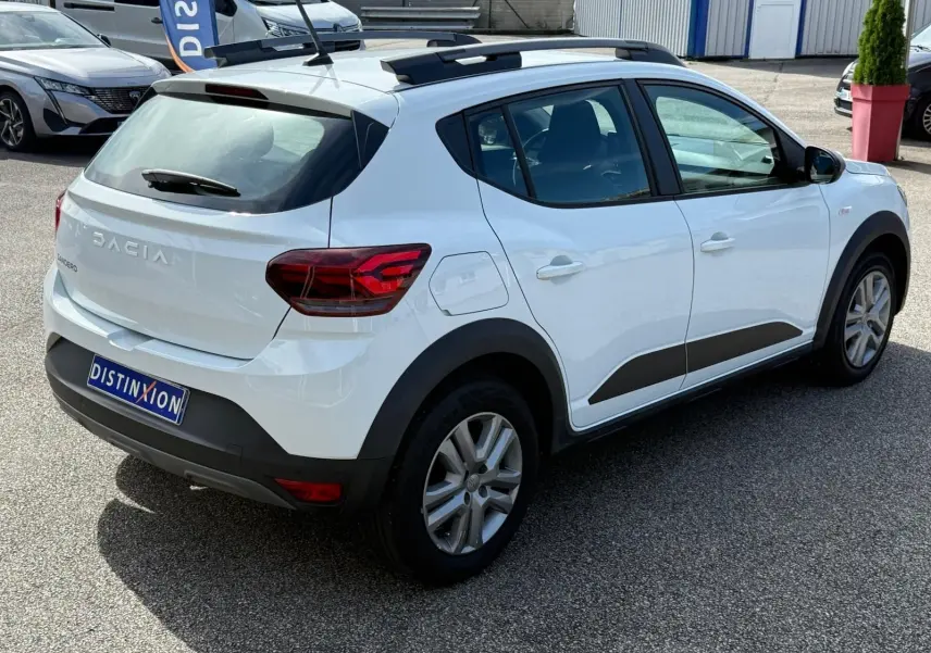 Vue 3/4 arrière droite d'une Dacia Sandero blanche 2023 avec protections noires et toit ouvrant visible.