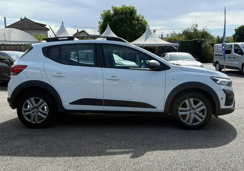 Profil côté gauche d'une Dacia Sandero blanche 2023 avec protections noires et barres de toit sur parking extérieur.