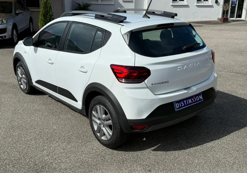 Vue 3/4 arrière droite d'une Dacia Sandero blanche 2023 avec toit ouvrant et protections noires sur les passages de roues.