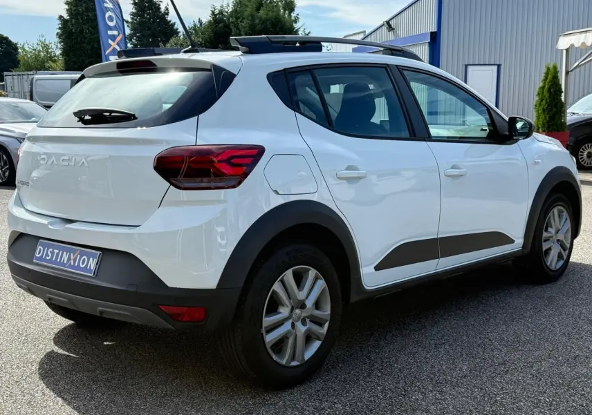 Vue 3/4 arrière droite d'une Dacia Sandero blanche 2023 avec protections noires et toit ouvrant visible.
