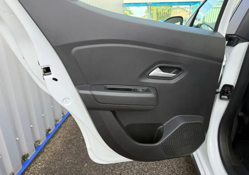 Porte arrière gauche ouverte d'une Dacia Sandero blanche 2023, montrant le panneau intérieur noir avec poignée et haut-parleur intégré.