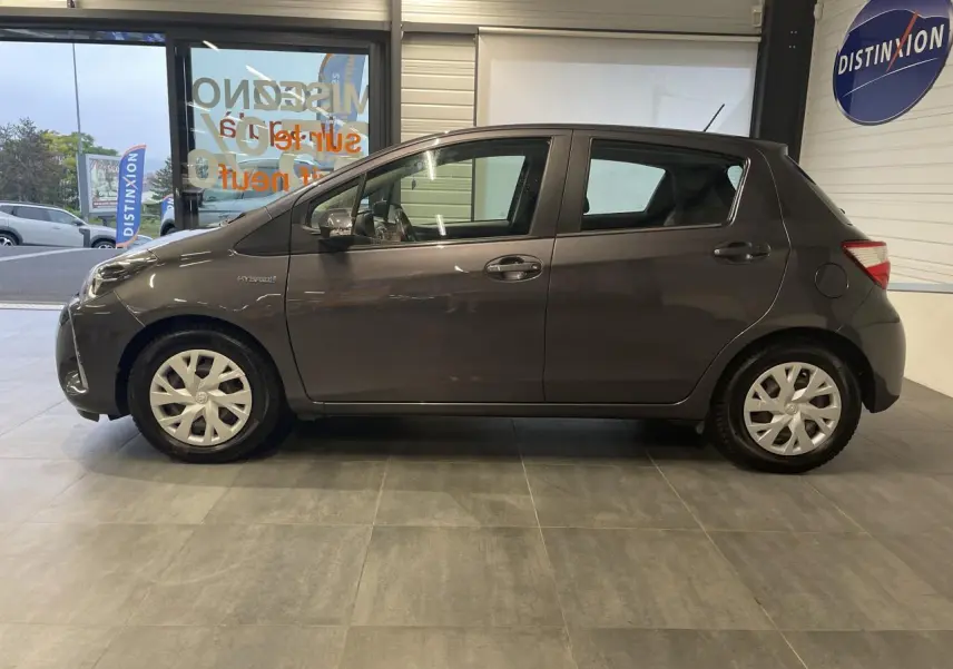 Profil côté gauche d'une Toyota Yaris hybride 2020 gris foncé métallisé avec enjoliveurs blancs en intérieur showroom.