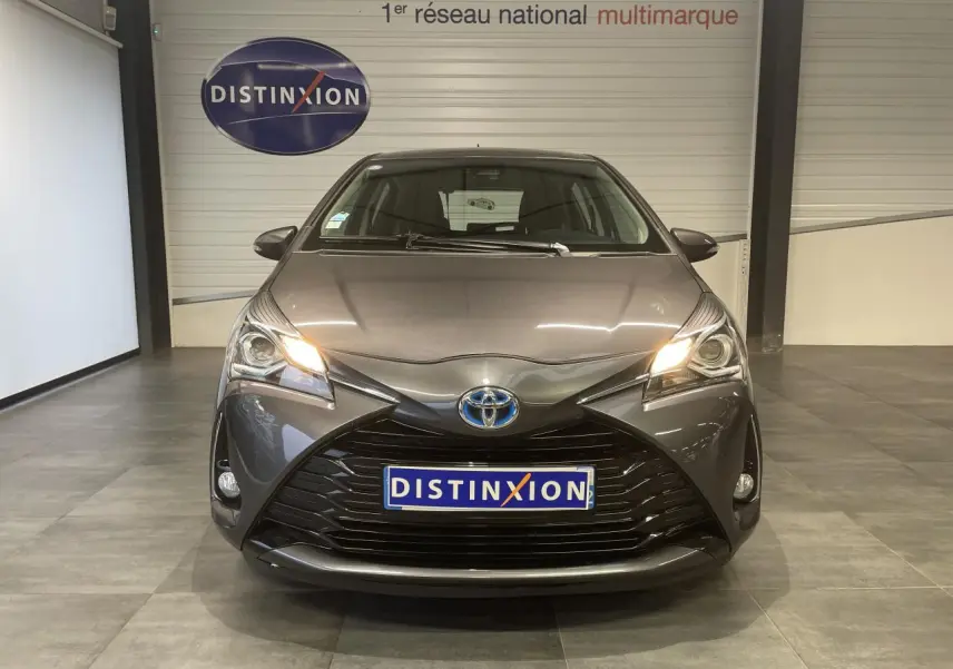 Vue frontale d'une Toyota Yaris hybride gris foncé métallisé avec calandre noir laqué et phares allumés.
