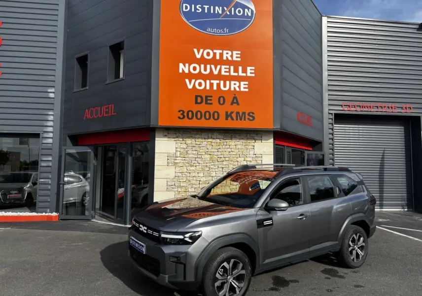 SUV Dacia Bigster gris foncé vu en 3/4 avant droit devant un bâtiment commercial avec enseigne orange.