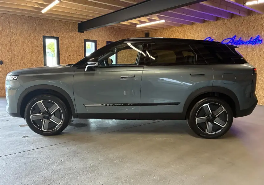 SUV JAECOO J7 2026 gris olive avec toit noir, vu de profil côté gauche dans un intérieur en bois.