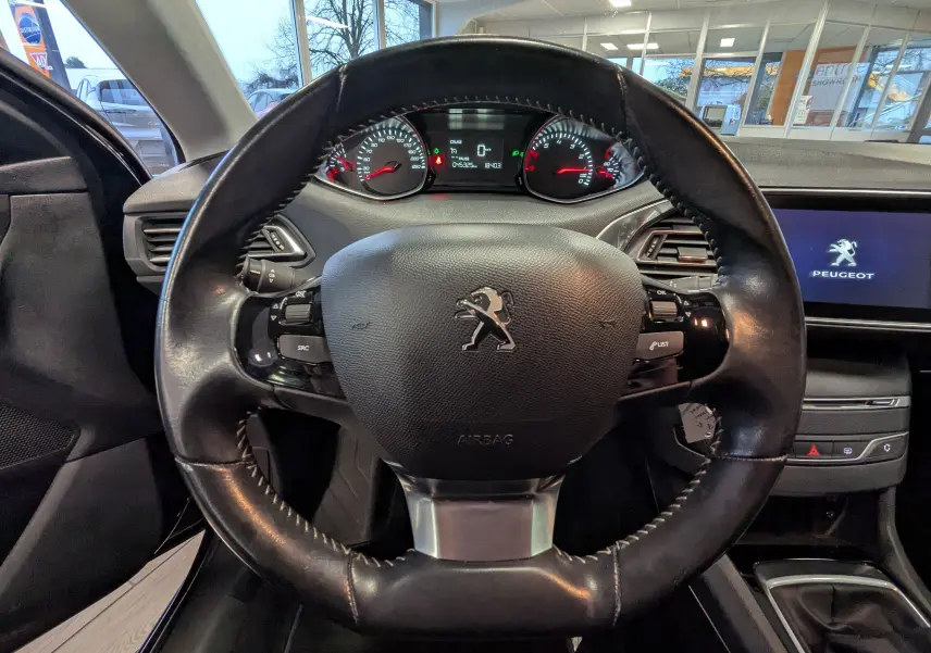 Vue intérieure centrée sur le volant cuir noir de la Peugeot 308 1.2 PureTech 110 Style avec tableau de bord et écran tactile allumé.