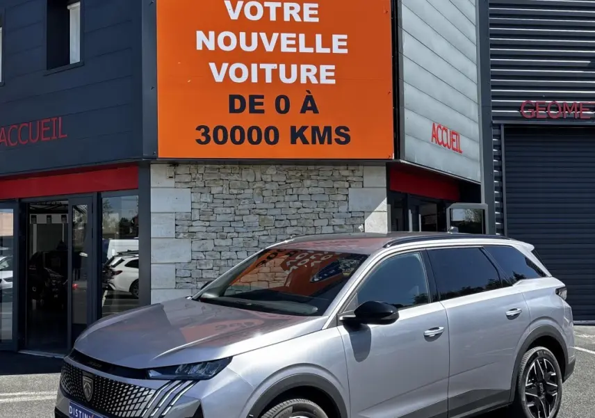 Peugeot 5008 gris clair en 3/4 avant droit, avec calandre distinctive et jantes noires, devant un bâtiment commercial.