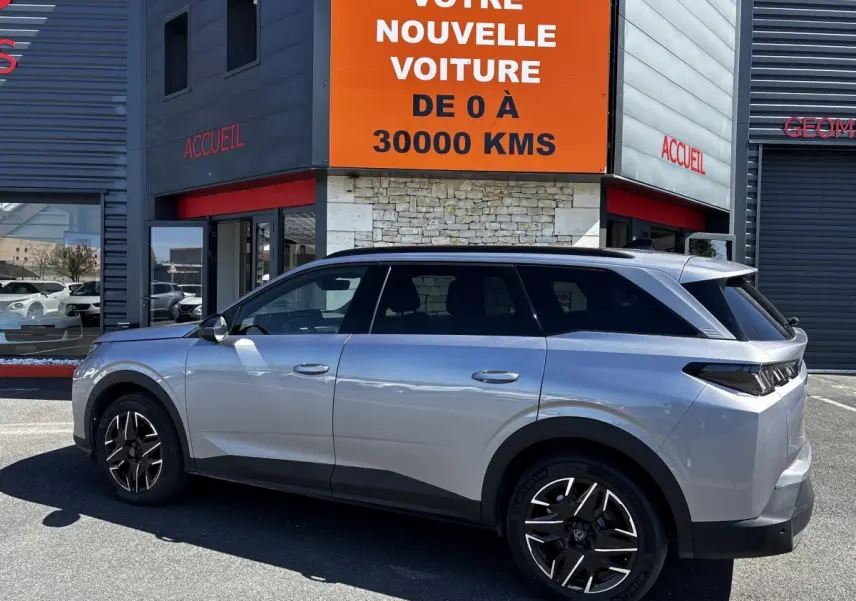 Vue de profil côté gauche d’un Peugeot 5008 gris clair 2025 garé devant un bâtiment avec panneau orange.