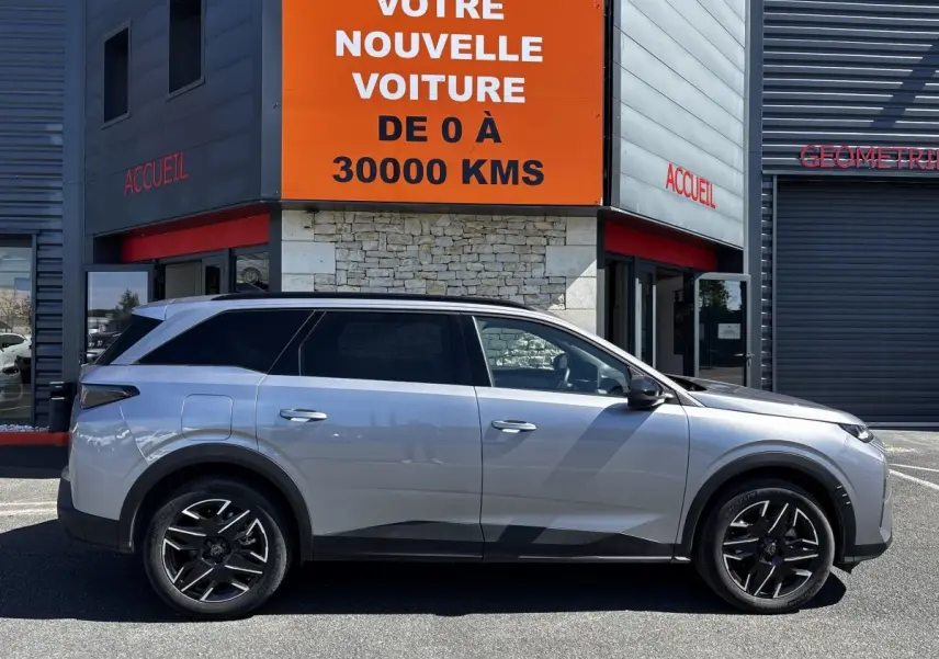 Peugeot 5008 gris clair vue de profil côté gauche, SUV hybride 2025 garé devant un bâtiment avec panneau orange.