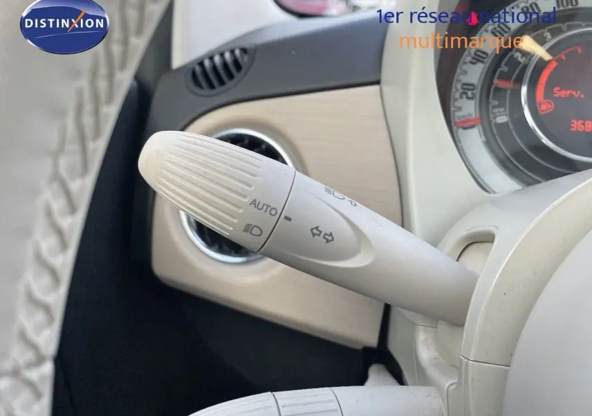 Gros plan sur la commande de clignotants blanche à gauche du volant d'une Fiat 500 blanche, tableau de bord partiellement visible.