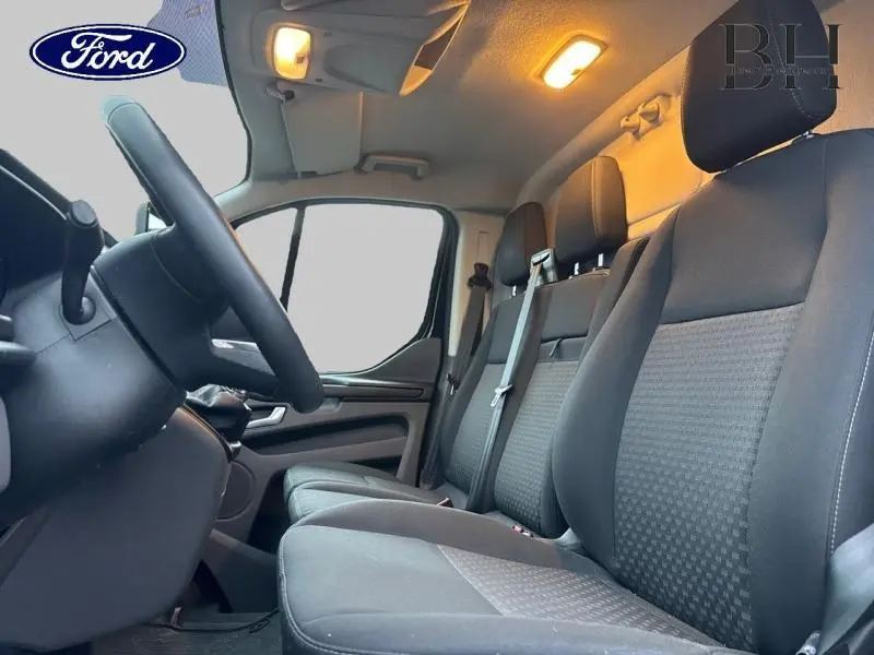 Vue intérieure côté conducteur du Ford Transit Custom Fourgon 2023, sièges tissu gris et volant cuir noir.