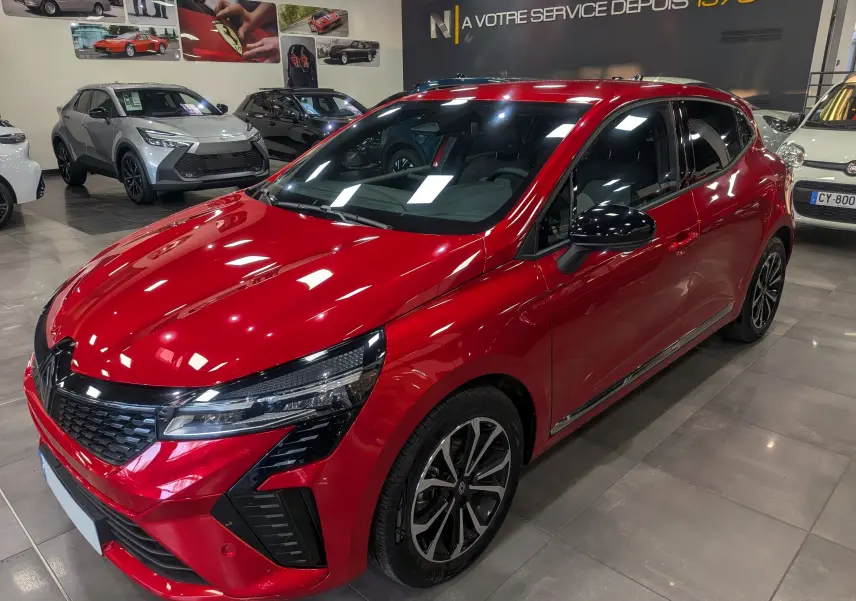 Renault Clio V Rouge Flamme vue 3/4 avant droit, avec jantes noires et détails noirs brillants sur calandre et rétroviseurs.